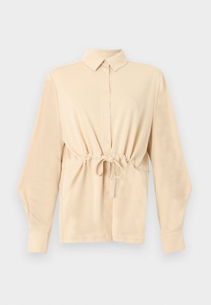 Chemise beige à manches longues avec col, boutons sur le devant et cordon de serrage à la taille, présentée sur un fond gris clair uni.