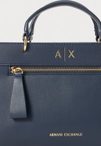 Sac à main en cuir bleu marine avec quincaillerie dorée, poche zippée et logo "A|X" en relief à l'avant. Fini texturé subtil.