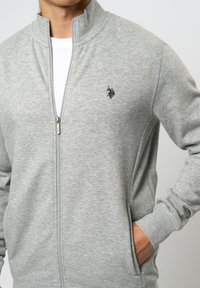Grå zip-up jakke laget av myk stoff, med høy krage, sidelommer og en liten brodert logo på brystet.