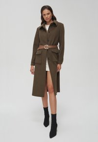 Oliwkowy płaszcz z kołnierzem, przednimi guzikami, dwoma dużymi kieszeniami i brązowym paskiem z kwadratową klamrą. Nosi się go na białej sukience.