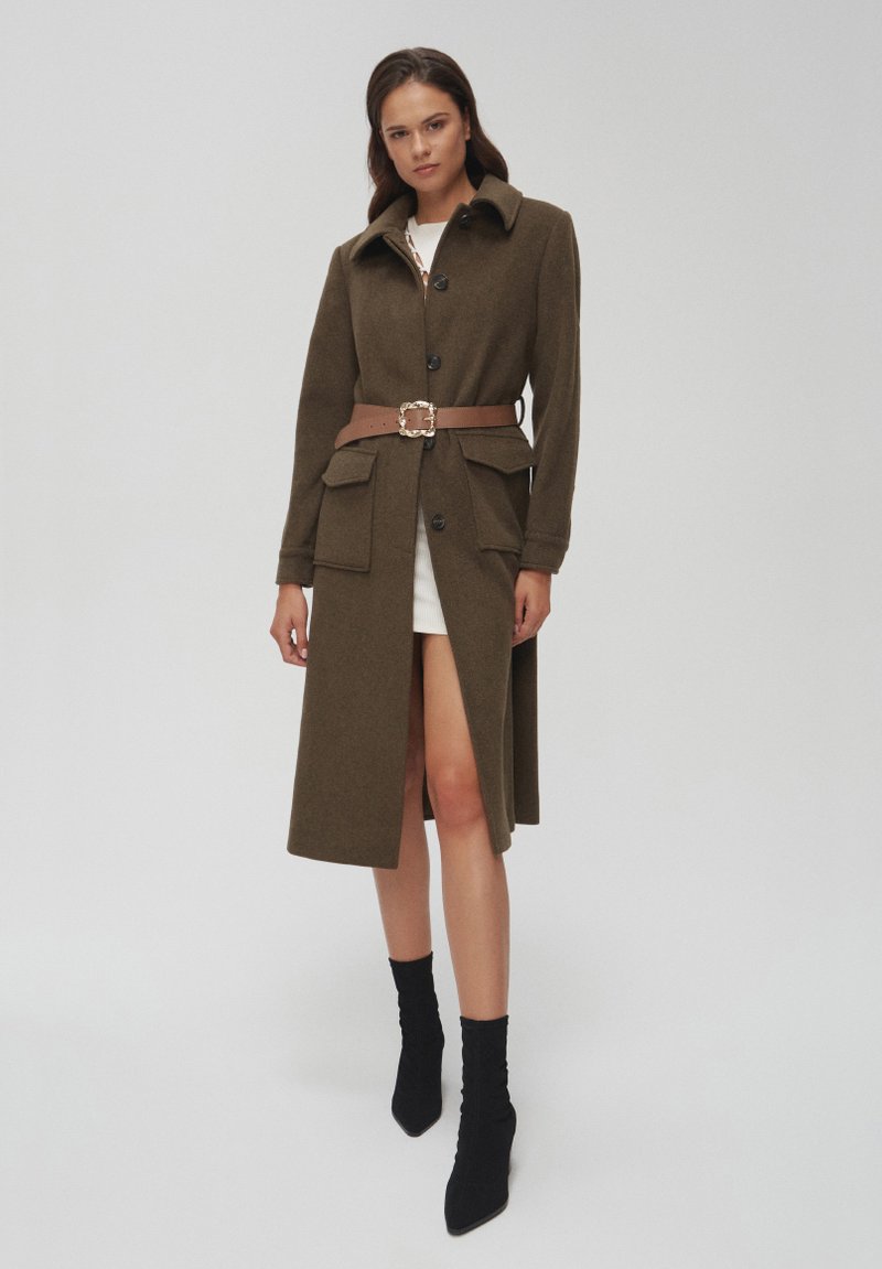 Oliwkowy płaszcz z kołnierzem, przednimi guzikami, dwoma dużymi kieszeniami i brązowym paskiem z kwadratową klamrą. Nosi się go na białej sukience.