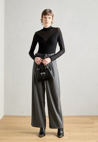 Top lungo trasparente nero con collo alto, abbinato a pantaloni grigi a gamba larga, una cintura nera e una piccola borsa nera. Stivali neri.