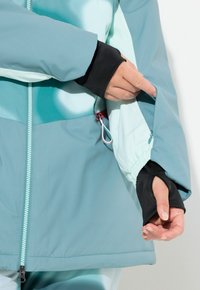 Světle modrá a tyrkysová nepromokavá bunda s kapsou na zip, elastickými manžetami a hladkou texturou. Detail zahrnuje kontrastní černé vnitřní manžety.