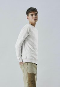 Hellgrauer Pullover mit langen Ärmeln und rundem Halsausschnitt, kombiniert mit beigen Hosen, die auf dem linken Bein einen strukturierten Cordura-Patch aufweisen.