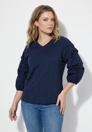 Kvinde med blond skulderlangt hår iført en navyblå bluse med opsamlede trekvart-ærmer og blå jeans, stående mod en hvid baggrund.