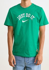 Green cotton T-shirt featuring "JUST DO IT" text in white arch and Nike logo in the center. Short sleeves, crew neck, relaxed fit.