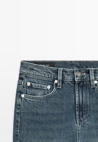 Donkere blauwe spijkerbroek met voorzakken, riemlussen, een knoopsluiting en zichtbare stiksels op een witte achtergrond.