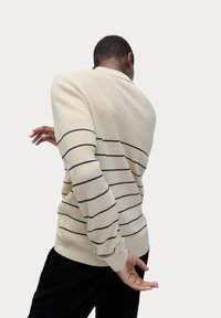 Beige strikket sweater med sorte horisontale striber, lange ærmer og en rund krave, båret vendt væk mod en lys baggrund.
