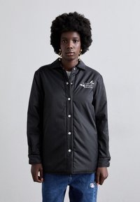 Zwarte button-up jas gemaakt van een glad materiaal, met een geborduurd ontwerp en drukknoopjes, gecombineerd met blauwe jeans.