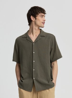 Giovane uomo con indosso una camicia verde oliva a maniche corte con bottoni e pantaloni beige, in piedi con le mani nelle tasche, guarda verso destra.