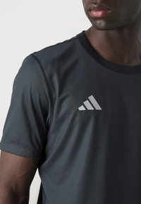 Mørk grå atletisk T-shirt lavet af glat stof. Har korte ærmer og en rund halsudskæring, med et logo med tre striber på forsiden.