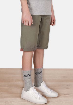 Helle olivgrüne Shorts mit umgeschlagenen Säumen, kombiniert mit grauen Socken mit schwarzem Text und weißen Sneakern mit perforierten Akzenten.