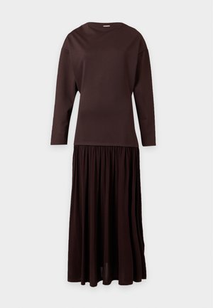 Mulher com cabelo curto e escuro, vestindo uma blusa castanha escura de manga comprida, saia comprida a condizer e sapatos planos pretos, em pé sobre um fundo simples.