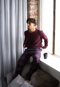 Maroonová košile s dlouhým rukávem a tmavě modré kárované pyžamové kalhoty. Jemná textura, volný střih. Tmavý keramický hrnek na okenním parapetu vedle polštářů.