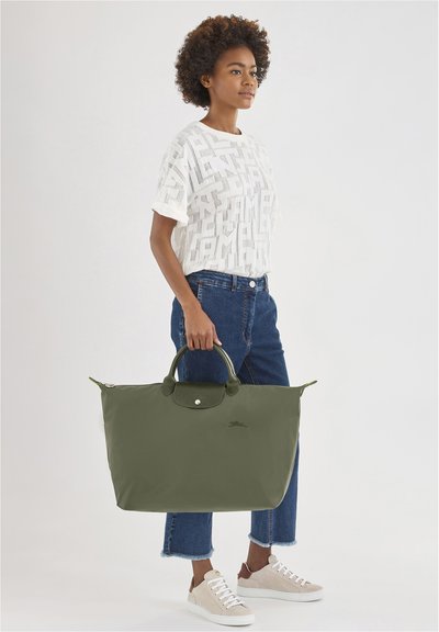 Green nylon tote bag with leather accents and dual handles, featuring a smooth texture and logo on the front, held by a person wearing casual attire.