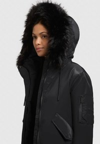 Black parka jacket with a large faux fur hood, front zip closure, side pockets, and a textured fabric finish. Features adjustable hood.