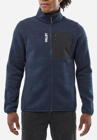 Veste en polaire bleu marine avec un col montant, fermeture éclair sur le devant, poche poitrine noire et poches latérales. Matériau texturé avec des motifs subtils.