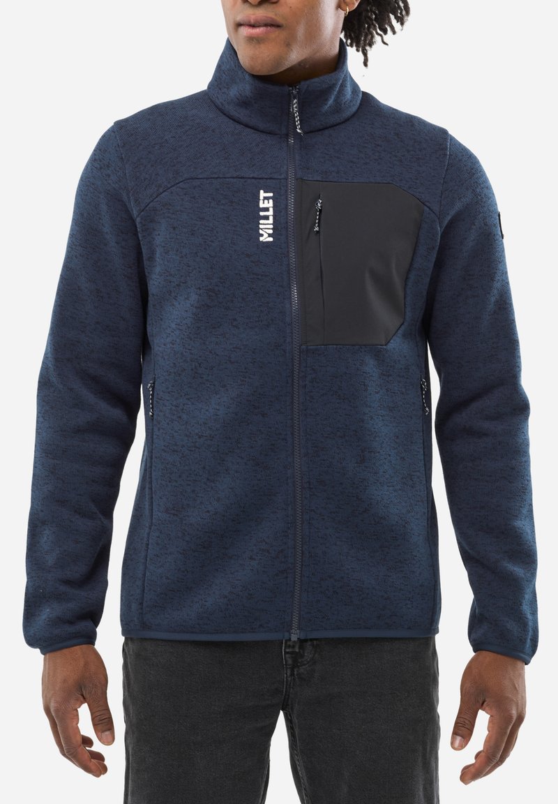 Veste en polaire bleu marine avec un col montant, fermeture éclair sur le devant, poche poitrine noire et poches latérales. Matériau texturé avec des motifs subtils.