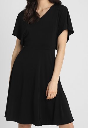 Femme portant une robe noire longueur genou avec des manches flottantes et un col en V, debout devant un fond uni gris clair.