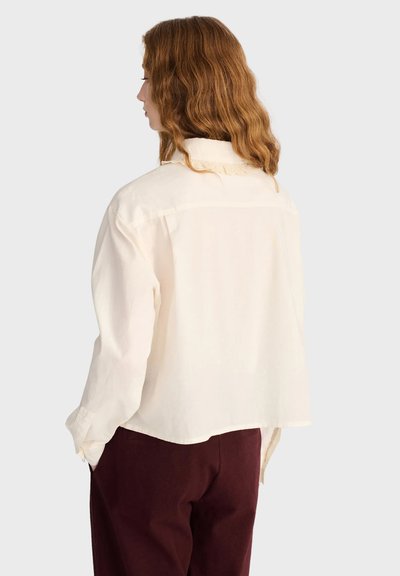 Femme aux cheveux roux ondulés mi-longs, de dos, portant une chemise blanche ample à manches longues et un pantalon bordeaux foncé.