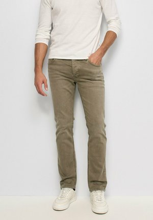 Hombre con pantalones ajustados beige, camisa blanca de manga larga y zapatillas blancas, de pie con una mano en el bolsillo.