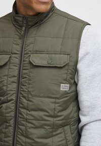 Olivgrüne Steppweste mit hohem Kragen, zwei Fronttaschen und einem Logopatch. Getragen über einem grauen Sweatshirt mit strukturiertem Finish.