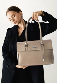 Bolso de cuero beige con diseño estructurado, asas superiores, detalles metálicos y etiqueta. Textura suave con acabado granulado.