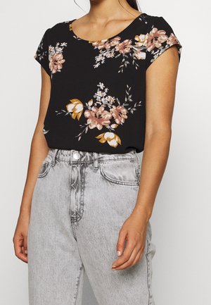 Femme portant un haut noir à manches courtes avec un imprimé floral rose et jaune, rentré dans un jean gris clair taille haute.