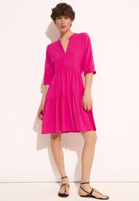 Fuksjowa sukienka z warstwowym wzorem, z półrękawami, dekoltem w serek i guzikami z przodu. Noszona z płaskimi czarnymi sandałami z złotym akcentem.