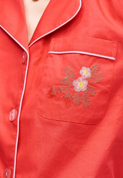 Rotes Hemd mit Knöpfen und weißen Paspeln, mit einer Tasche, die rosa Blumen und rote Erdbeeren bestickt hat.