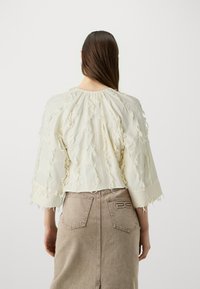 Light cream textured top with frayed accents, wide sleeves, and gathered neckline, paired with a taupe denim skirt featuring subtle branding.