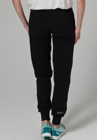 Pantalones de chándal negros con puños acanalados, un corte relajado y un detalle de logo cerca del tobillo. Llevados con zapatillas deportivas en color teal.
