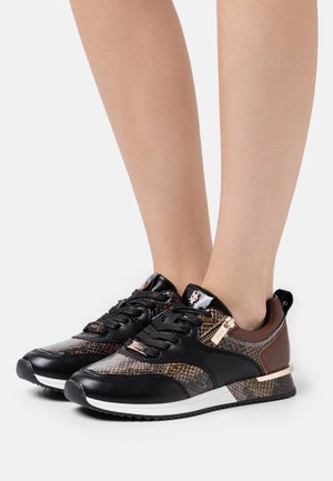 Stopy w czarno-brązowych trampkach z wzorem skóry węża, czarnymi sznurowadłami i białymi podeszwami na białym tle.