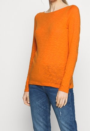 Femme portant un haut à manches longues orange vif et un jean bleu, debout devant un fond clair uni