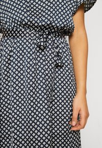 Robe bleu marine avec motif losange blanc, manches courtes et taille nouée. Fabriquée en tissu léger, elle présente des plis et une coupe décontractée.