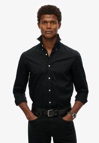Camisa negra de botones con collar estructurado, con botones blancos y un logo sutil. Combinada con un cinturón negro y un reloj de pulsera.