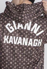Sudadera marrón con un patrón repetido de logotipos, con texto blanco que dice "GIANNI KAVANAGH" en la parte posterior. Tejido suave y texturizado.