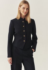 Blonde woman wearing a tailored black jacket with gold buttons and matching black pants, standing with one hand in pocket.