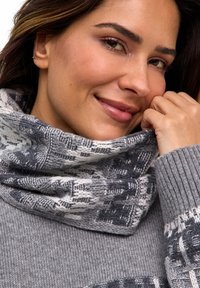 Donna sorridente, che indossa un maglione a maglia grigio e bianco con un motivo e un caldo collo abbinato, tenendo il collo vicino al viso.