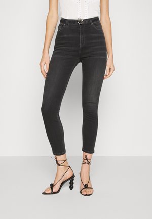 Femme portant un jean skinny noir taille haute avec une ceinture noire et des sandales à talons hauts fines noires avec des talons sphériques.