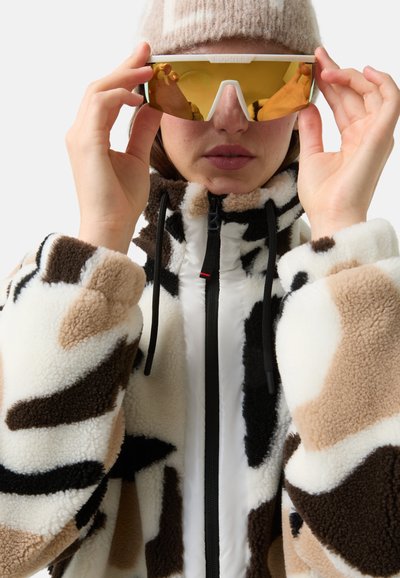 Fleecejack met een mix van crème-, bruin- en zwarte patronen. Voorzien van een hoge kraag en ritssluiting, gedragen met gele spiegelzonnebril.