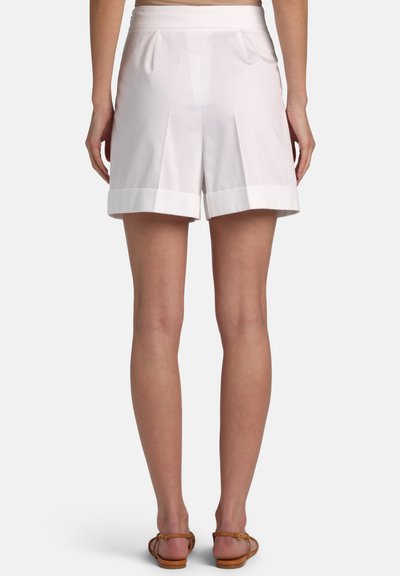 Hvide skræddersyede shorts med plisseringer og opbukket kant båret af en person, der står, kombineret med brune flade sandaler.