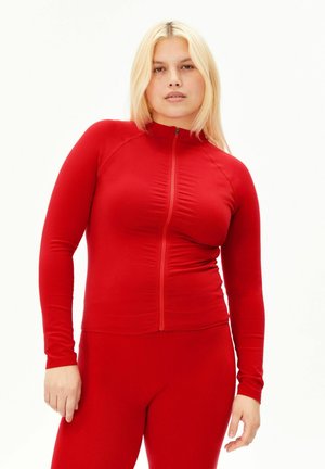 Junge Frau mit blonden Haaren, die ein eng anliegendes rotes langärmeliges Top mit Reißverschluss und passende rote Hose trägt, steht vor einem weißen Hintergrund.