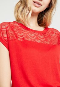 Donna che indossa un top rosso con maniche a cappuccio e uno scollo in pizzo trasparente con un motivo a onde, mostrata dalle spalle alle labbra.
