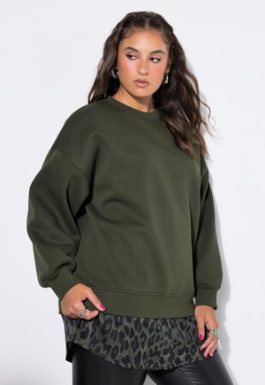 Ung kvinde med langt brunt hår iført en oversized mørkegrøn sweatshirt over en leopardmønstret skjorte og sorte bukser, der kigger sidelæns.