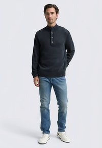 Mörk marinblå stickad tröja med ribbad krage, tre knappar i knappslå och texturerat mönster, kombinerad med ljusblå jeans och vita sneakers.