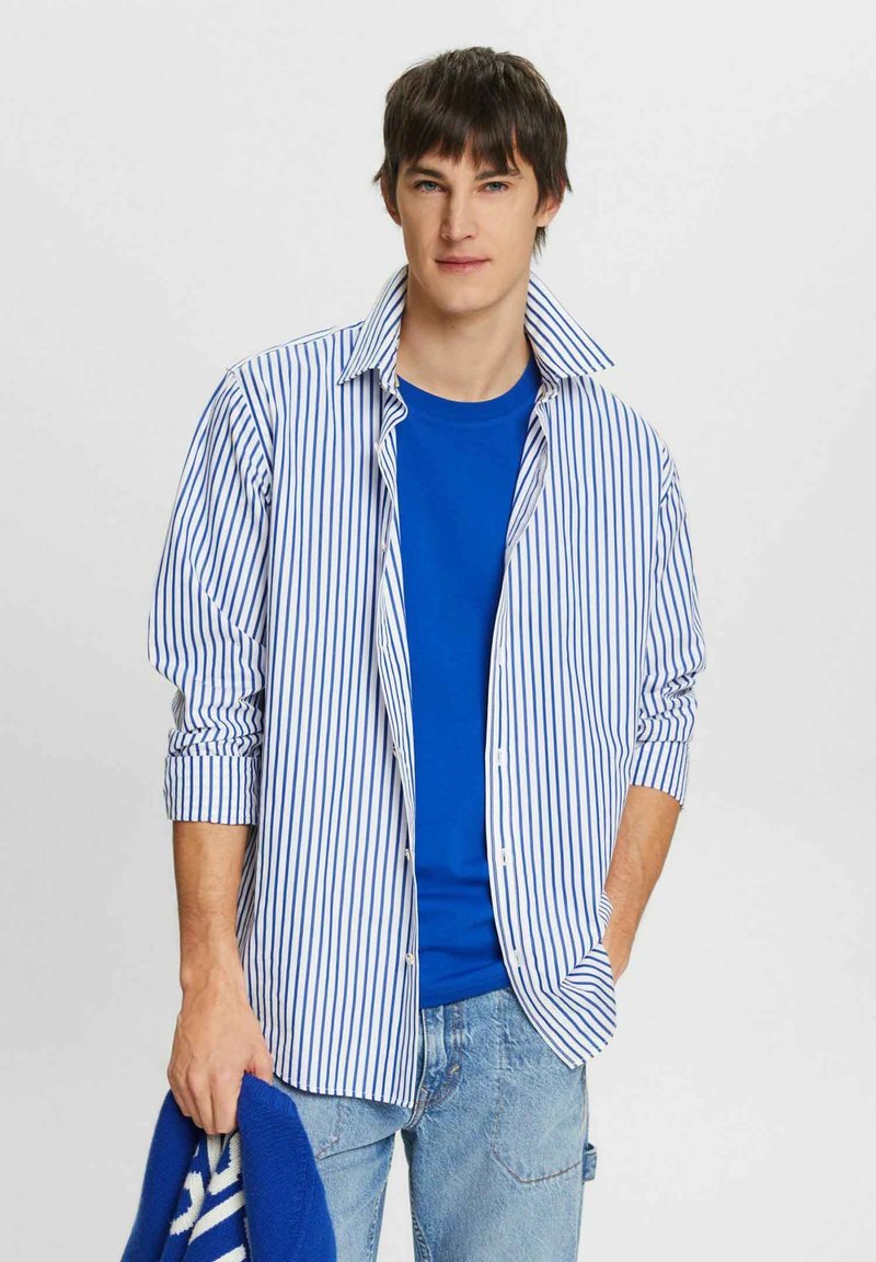 Vit skjorta med blå vertikala ränder, lagrad över en blå t-shirt. Ljusblå jeans och håller en blå tröja. Avslappnad passform, knäppning framtill.