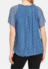 Modrá blůza s potiskem a krátkými, průsvitnými rukávy. Má nařasený výstřih a geometrické tvary v různých odstínech modré. Lehká látka.