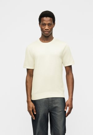Jeune homme debout face à l'avant, portant une chemise à manches courtes unie de couleur blanc cassé et un pantalon gris foncé, sur un fond clair.