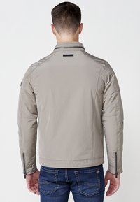 Chaqueta acolchada gris claro con cuello alzado, bolsillos con cremallera en los laterales y detalles de diseño deportivo en los hombros y las mangas. Llevada sobre jeans azules.